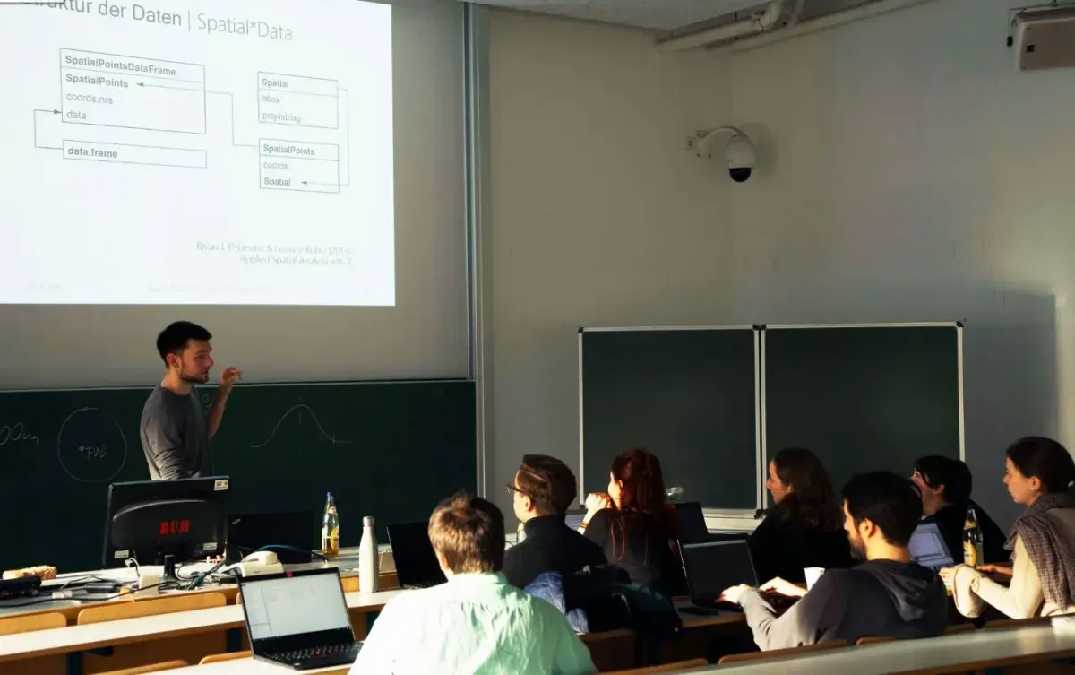 Bild eines Seminarraums. Eine Person steht vorne und erklärt einer Gruppe von ca. 10 sitzenden Personen etwas. Im Hintergrund die an die Leinwand geworfenen Folien. Alle Personen haben einen Laptop vor sich. 