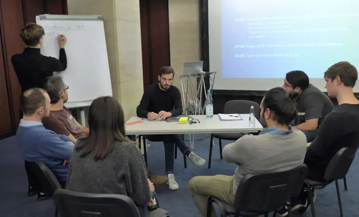 Eine Gruppe von Personen, die zusammenarbeiten und diskutieren. Eine Person sitzt an einem weißen Tisch mit 6 Personen, die in einem Halbkreis um den Tisch herum sitzen. Eine Person im Hintergrund steht und macht sich Notizen auf einem Flipchart