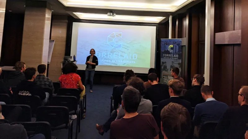 Foto eines Raums mit etwa 20 Personen, die in Reihen sitzen. Vor den Stuhlreihen steht Sylvi, die gerade einen Vortrag hält. Hinter ihr ist auf einer Leinwand die Präsentationsfolie zu sehen, die sie gerade präsentiert. Sie zeigt das CorrelAid-Logo.