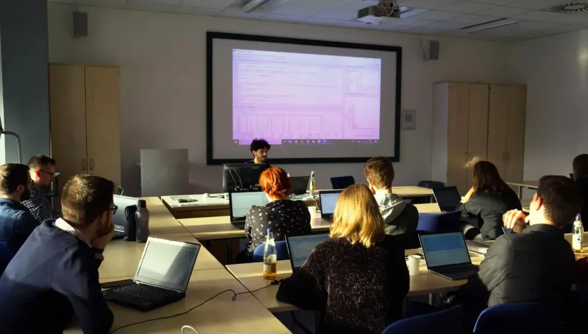 Bild eines Seminarraums in einer Universität. Ca. 20 Personen sitzen an Tischen und hören der vortragenden Person zu, die vorne sitzt. Auf der Leinwand hinter dieser Person ist der geteilte Bildschirm der Person zu sehen. RStudio ist geöffnet. Die meisten Personen haben Laptops vor sich. 