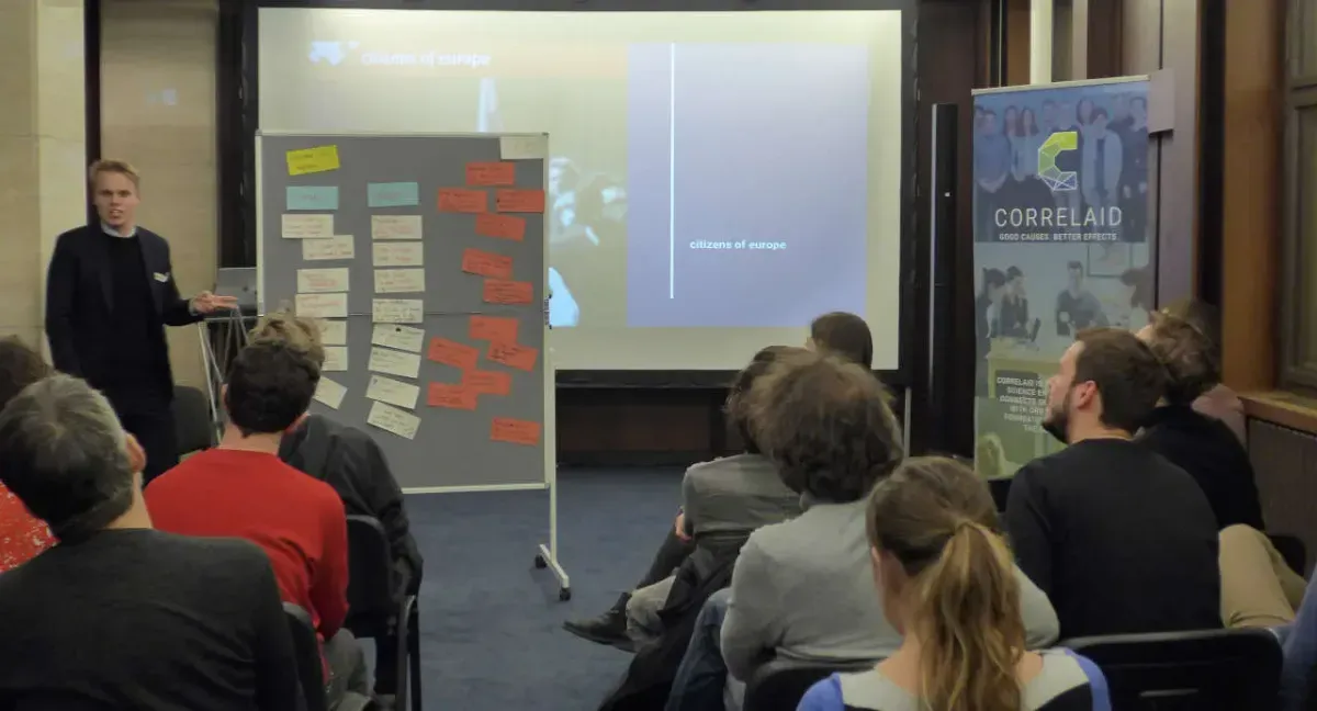Eine Person präsentiert eine Reihe von Post-its, die an eine Tafel geheftet sind. Eine Gruppe von etwa 10 Personen sitzt in Stuhlreihen und hört aufmerksam zu. Im Hintergrund ist ein Roll-up zu sehen, das für CorrelAid wirbt.